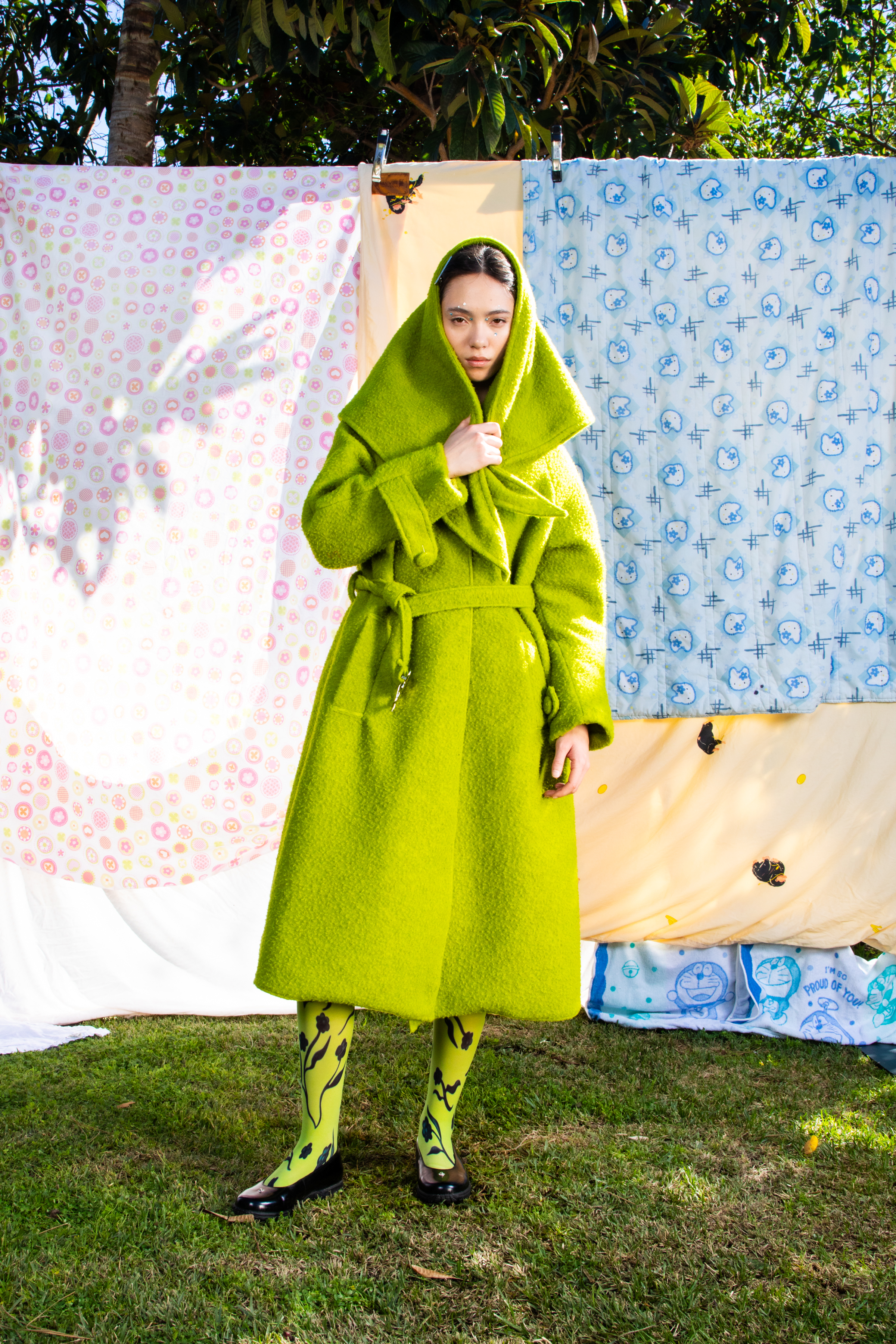 Full body image of a female model in a green matching coat and scarf set against a backdrop of patterned quilts.